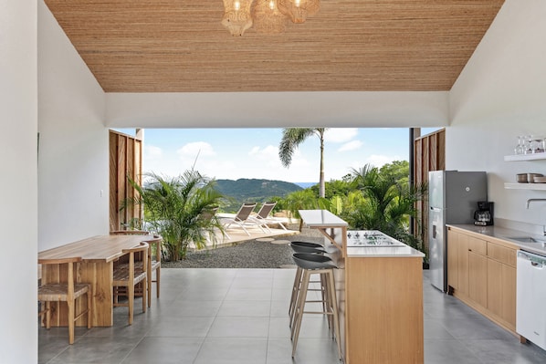 Cocina moderna con barra, mesa de comedor y sillas. Se ve un patio con sillas y plantas.