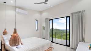 A bedroom with a large bed, a ceiling fan, and a view of a green landscape through a sliding glass door.