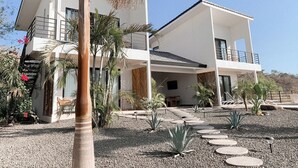 A modern two-story house with a balcony, surrounded by a gravel driveway and tropical plants.