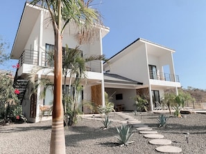 Una casa moderna de dos pisos con un balcón, rodeada por un camino de grava y plantas tropicales.