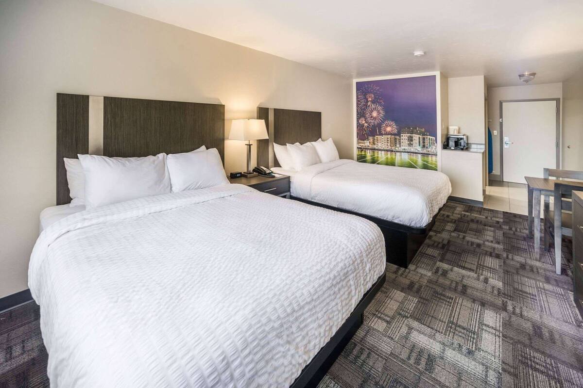 Une chambre d’hôtel avec deux lits, un bureau, une chaise, une table de chevet, une lampe et une fresque représentant un paysage urbain sur le mur.