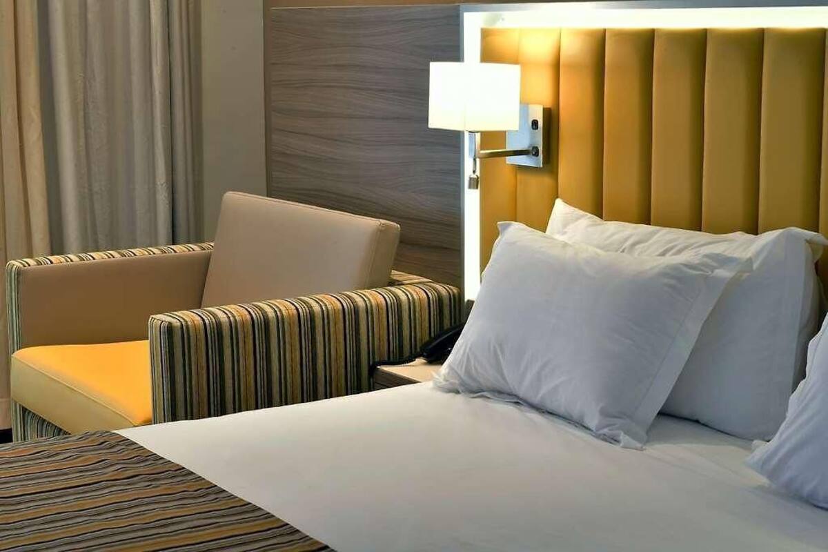 Quarto de hotel com cama, poltrona listrada, mesa de cabeceira e luminária fixa na parede.