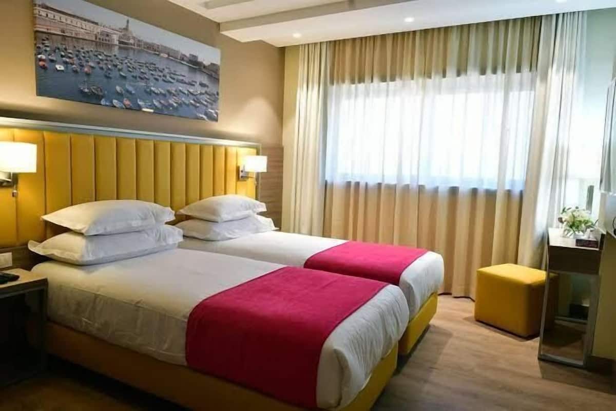 Quarto de hotel com duas camas, uma escrivaninha e uma janela com cortinas.