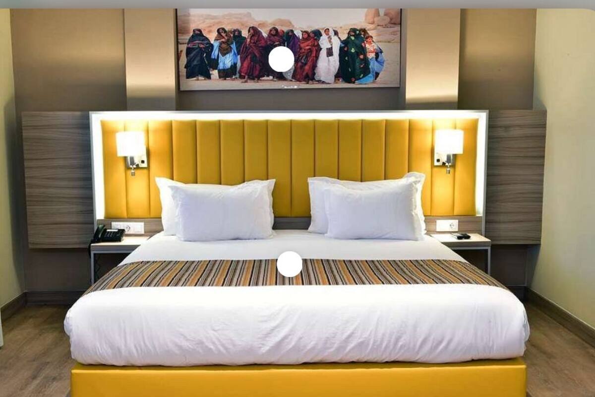 Quarto de hotel com uma cama grande, duas mesas de cabeceira, cabeceira com iluminação integrada e uma obra de arte pendurada na parede sobre a cama.