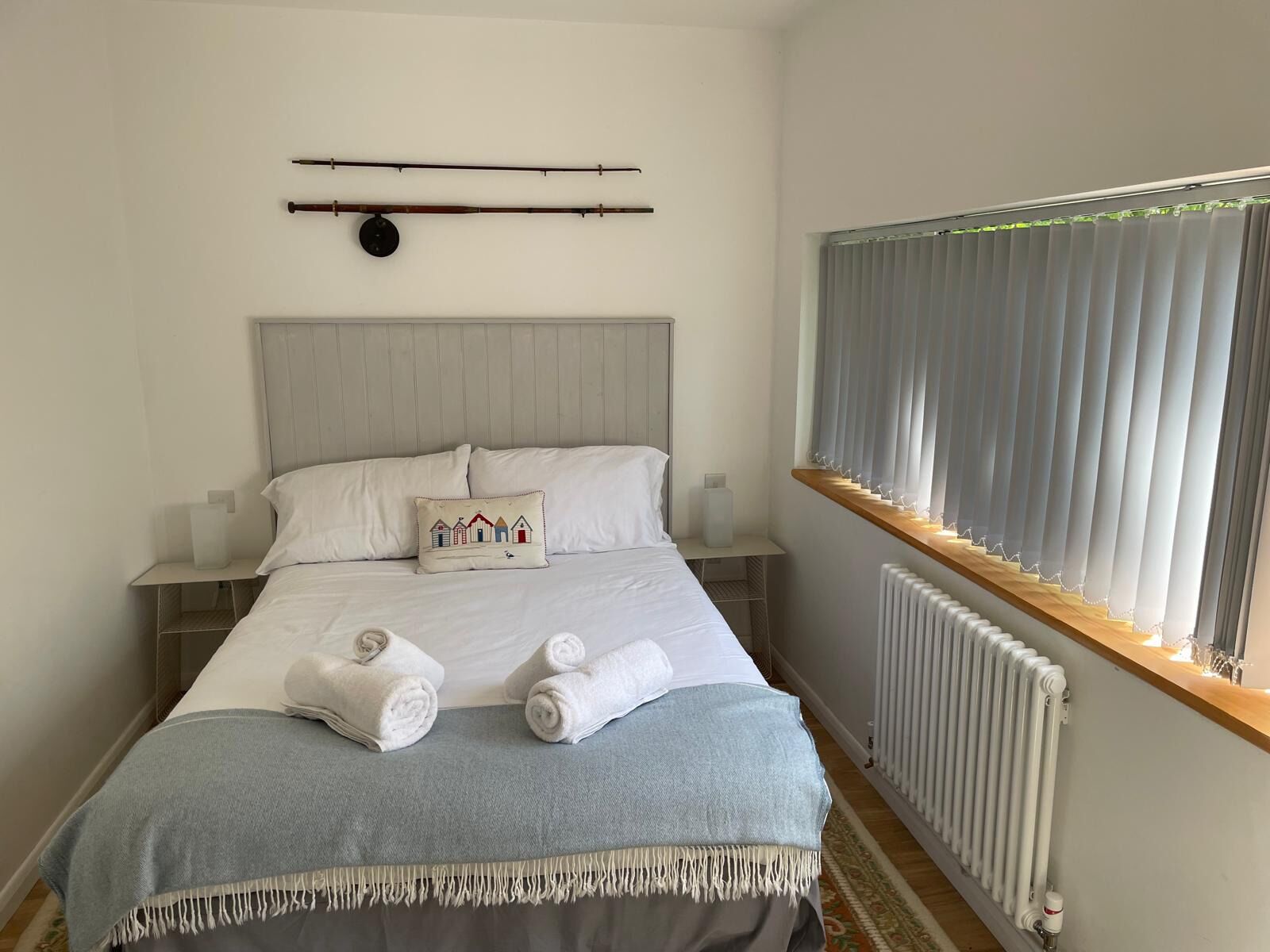 A neatly made bed with pillows, a bedside table, a wall-mounted clock, a window with blinds, and a radiator.