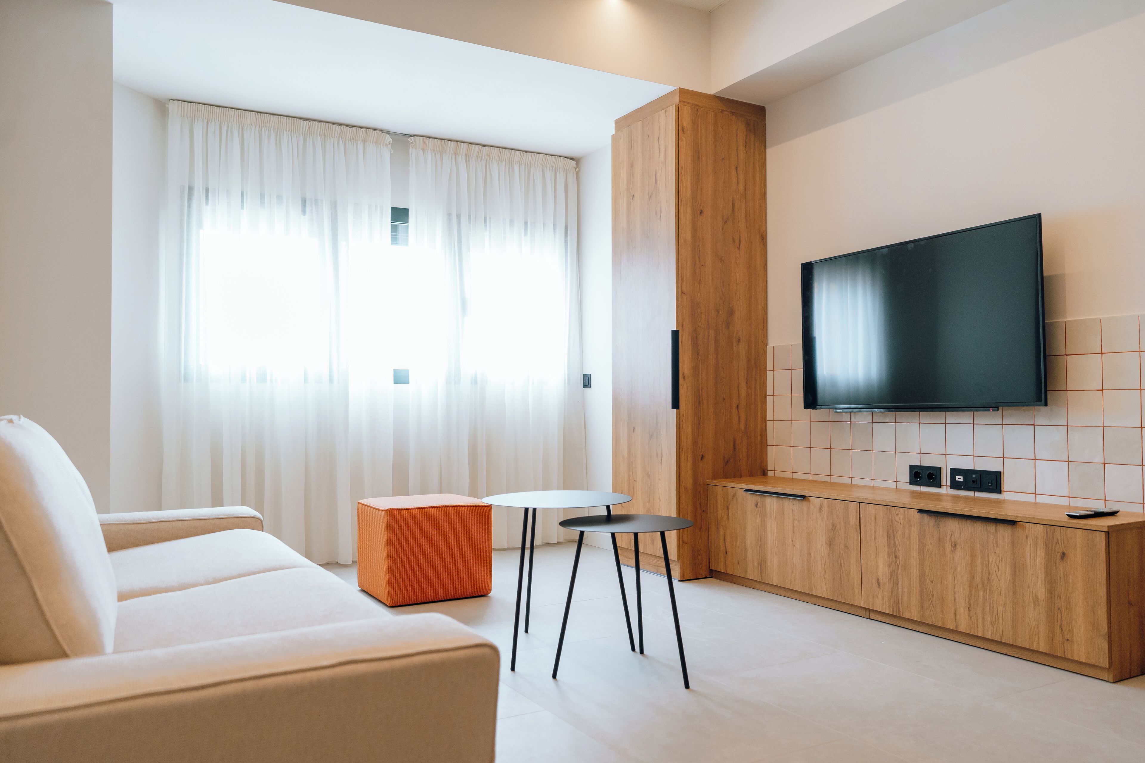 Una sala de estar moderna con un sofá, un pequeño taburete naranja, dos mesas redondas, un televisor montado en una pared de madera y un ventanal con cortinas translúcidas.