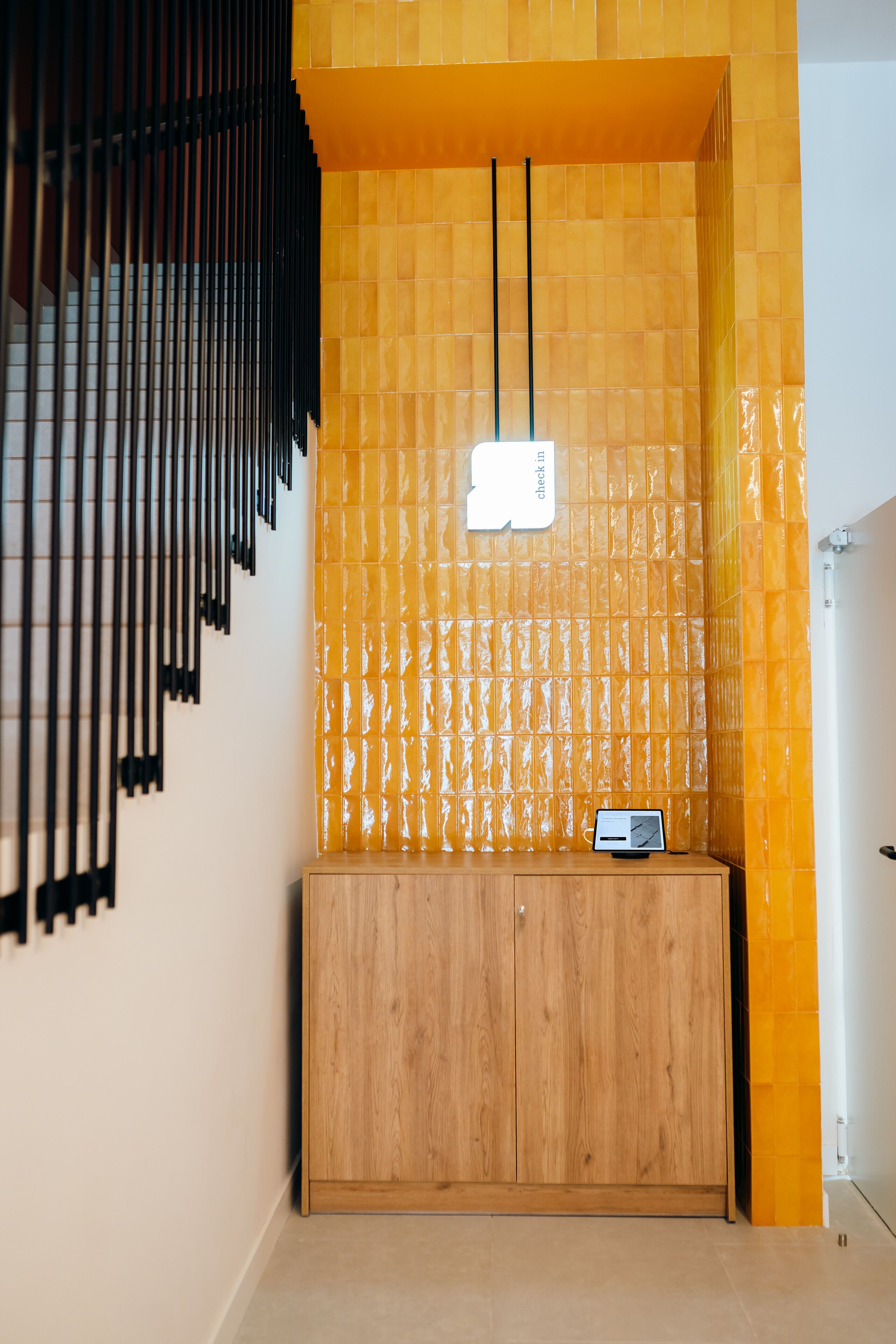 A wooden cabinet with a digital device on top, set against a wall with yellow tiles and black vertical elements.