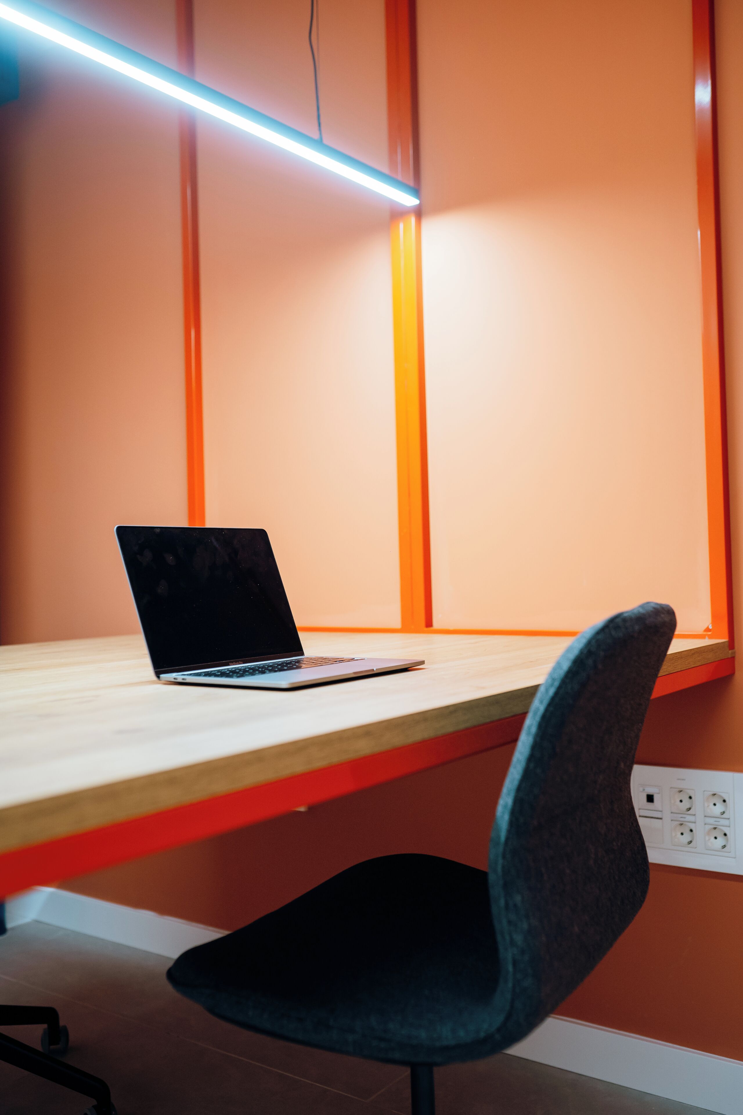 A workspace with a laptop on a wooden desk, a black chair, and orange walls.