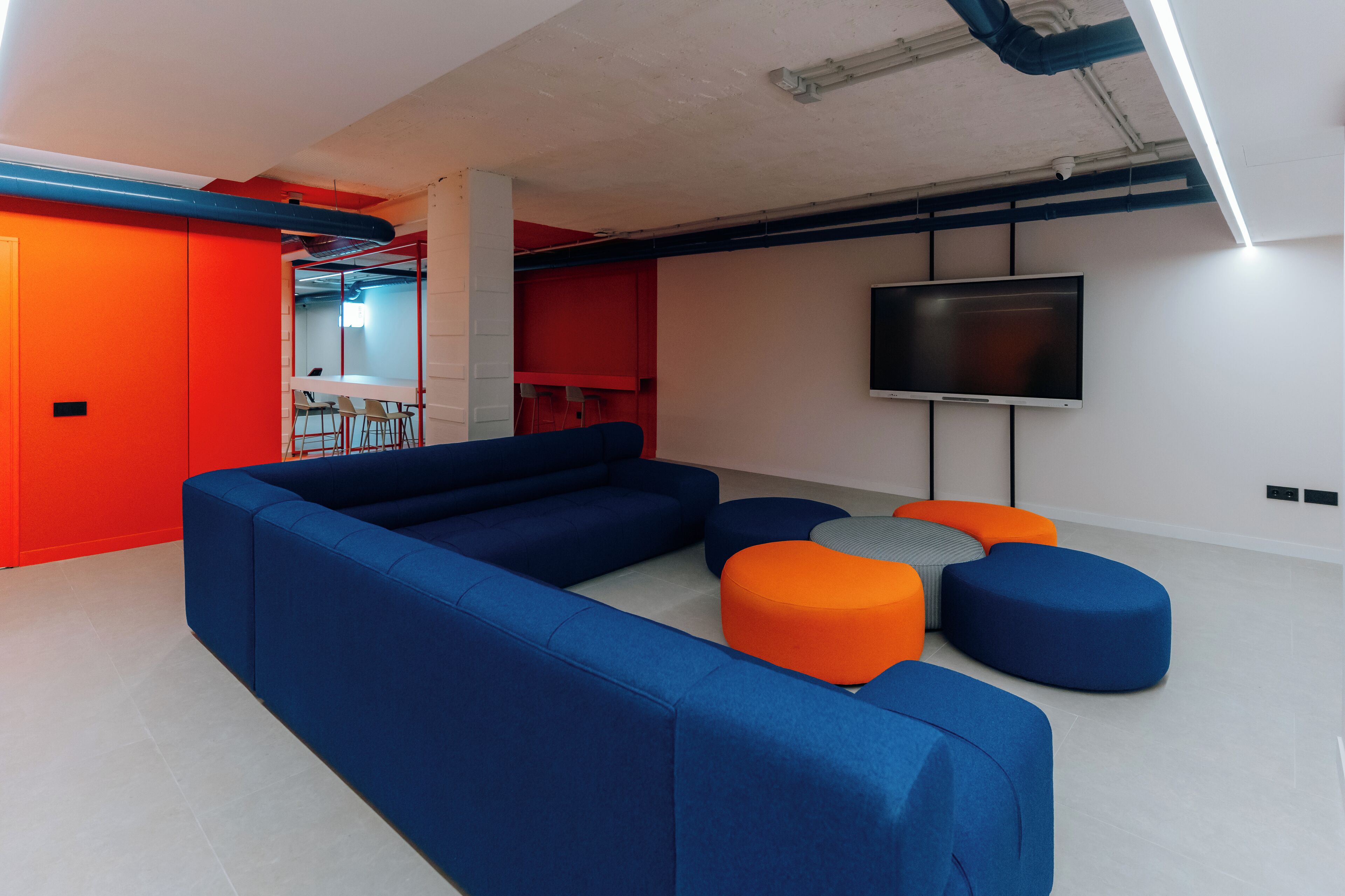 A modern living room with a sectional sofa, orange and blue ottomans, and a flat-screen TV mounted on the wall.
