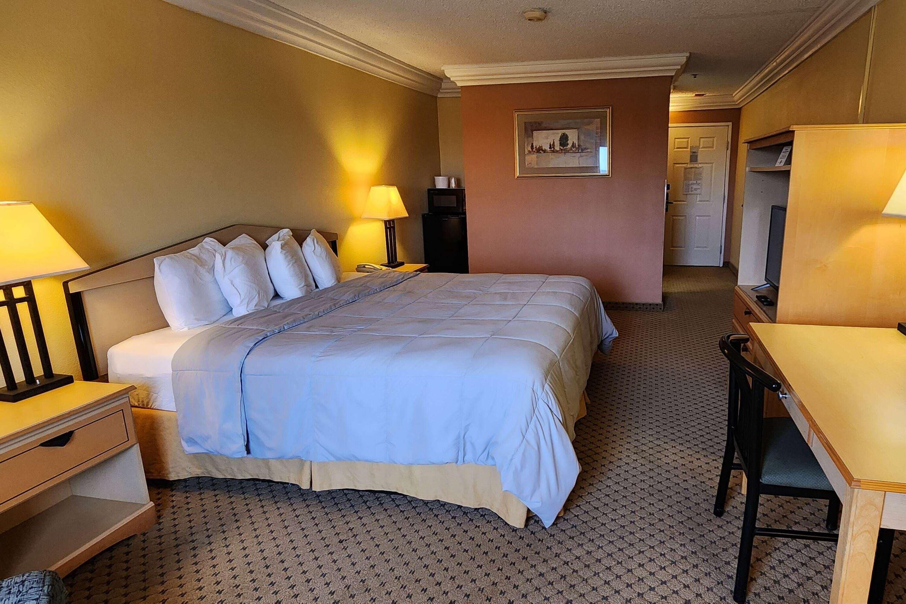 A hotel room with a large bed, two bedside tables with lamps, a desk with a chair, and a door leading to a bathroom.