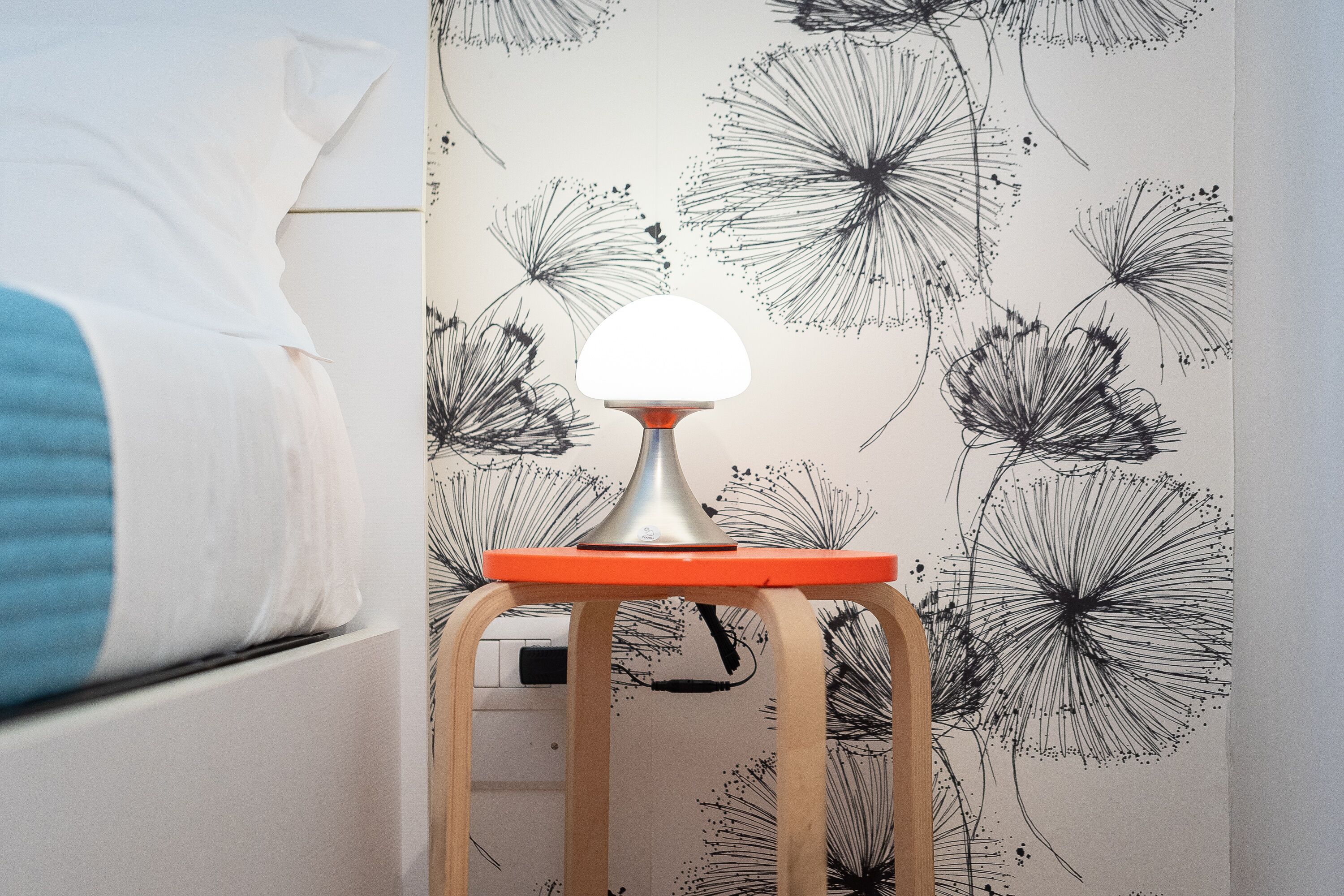 Una habitación con una cama, un taburete de madera con una lámpara de mesa, y una pared con un estampado de diente de león.