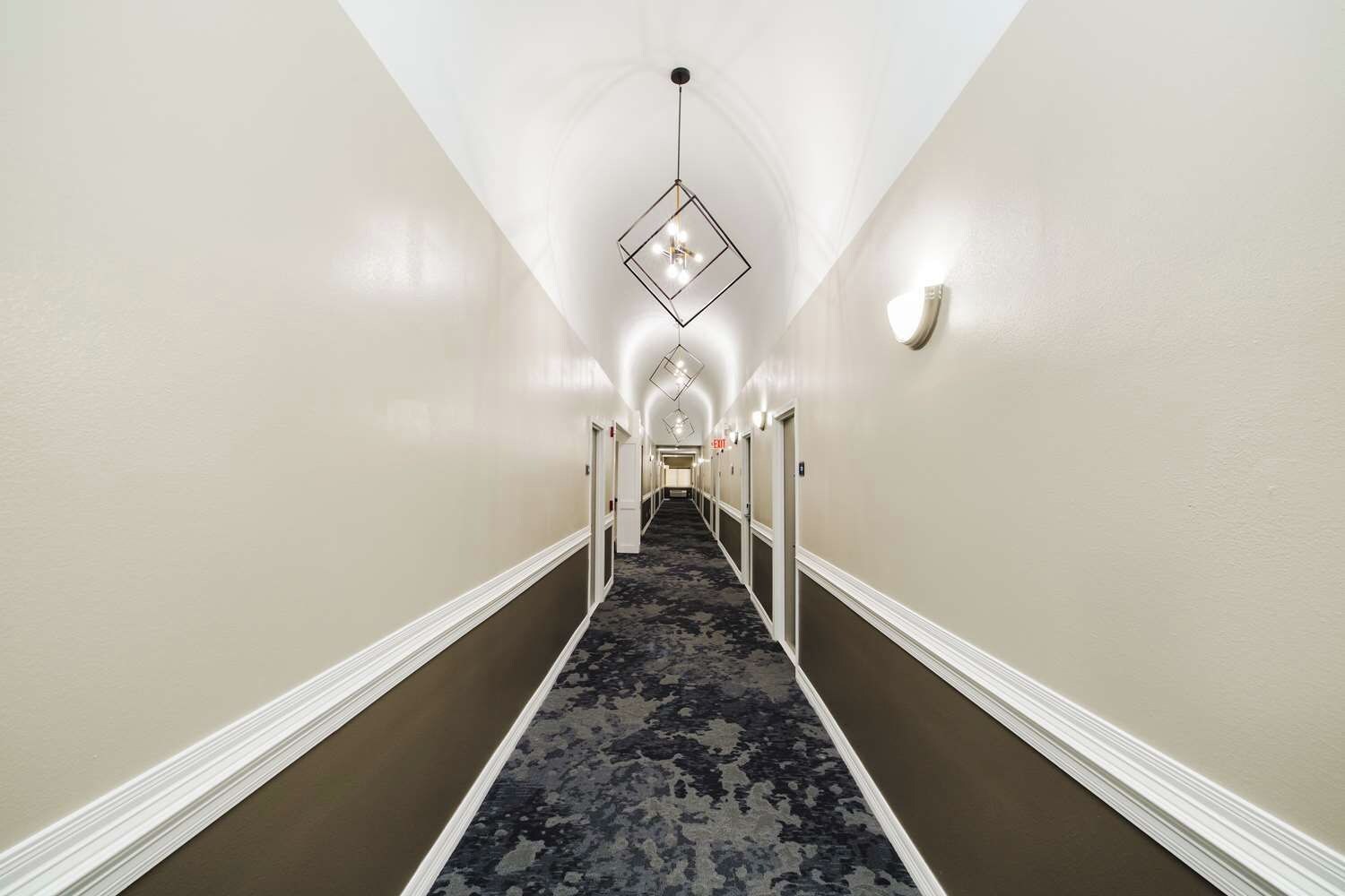 A hotel hallway with a patterned carpet, white walls, and a geometric light fixture.