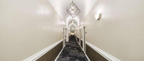 A hotel hallway with a carpeted floor, white walls, and a geometric light fixture.