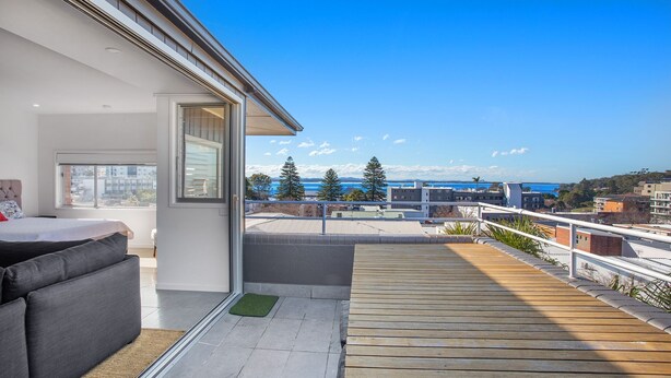 A modern balcony with a wooden deck, a grey sofa, and a view of the sea and buildings.
