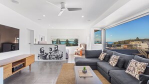 A modern living room with a grey sofa, a coffee table, and a flat-screen TV. There is a large window with a view of the city and a balcony.