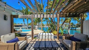 Uma área de estar externa com móveis de vime, uma mesa de madeira e vista para uma piscina e palmeiras.