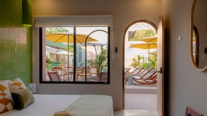 Hotelkamer met een bed, een hoed op het bed, een raam met uitzicht op een terras met parasols.