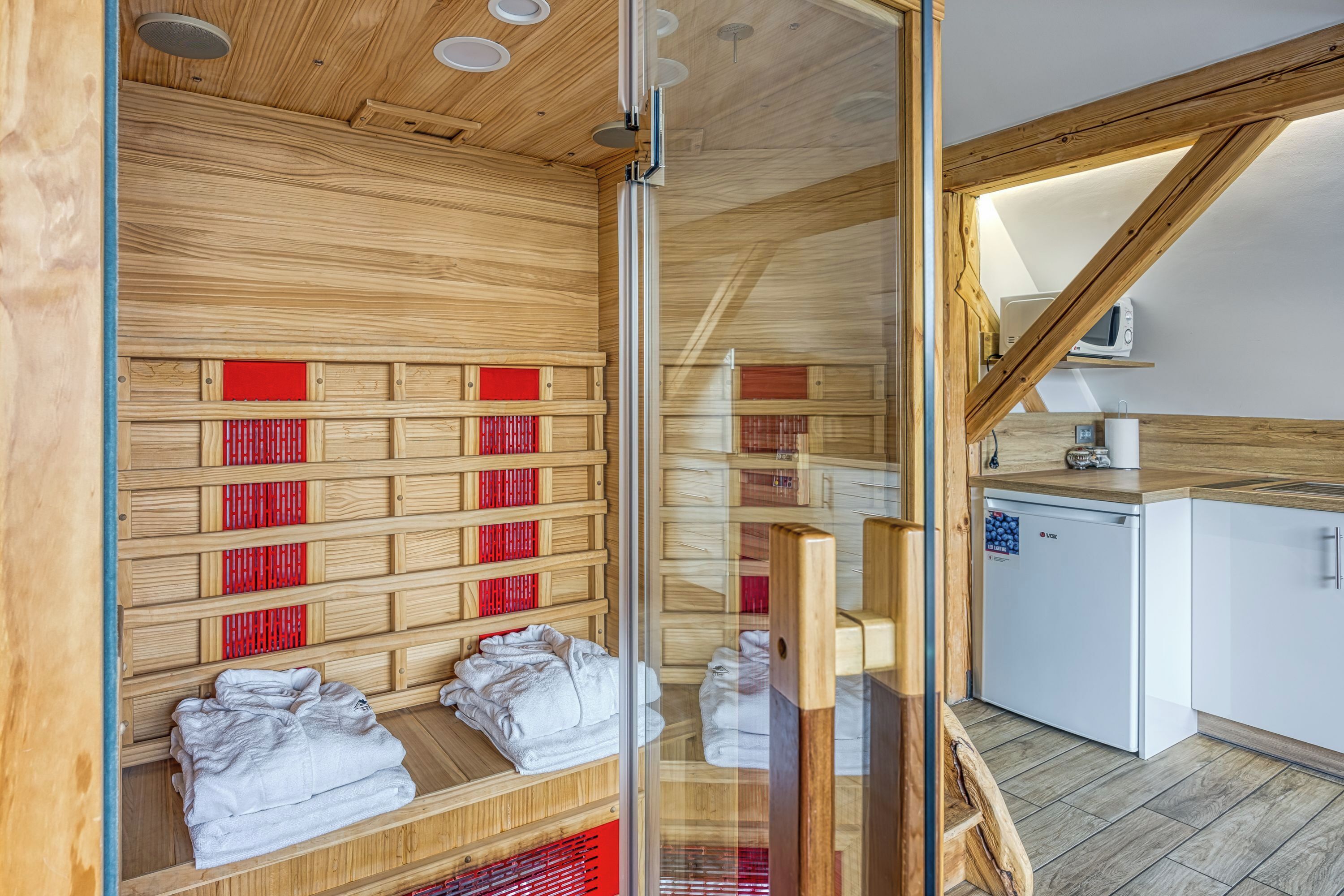 A sauna room with wooden benches and red heating elements, a glass door, and a white refrigerator in the adjacent kitchen area.