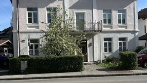 Un edificio de dos pisos con un balcón, un pequeño jardín y un cielo despejado.