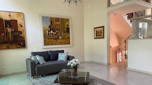Sala de estar com sofá, mesa de centro e tapete. Há quadros nas paredes e uma escada ao fundo.
