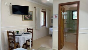 Quarto de hotel com área de jantar, TV fixada na parede, banheiro com chuveiro de vidro e porta de madeira.