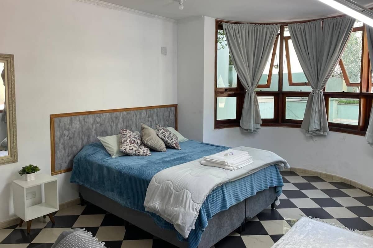 Quarto com piso xadrez, cama com colcha azul e branca, cabeceira e janela com cortinas.