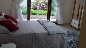 Quarto com cama, cortinas brancas, vista para um jardim e cômoda branca.