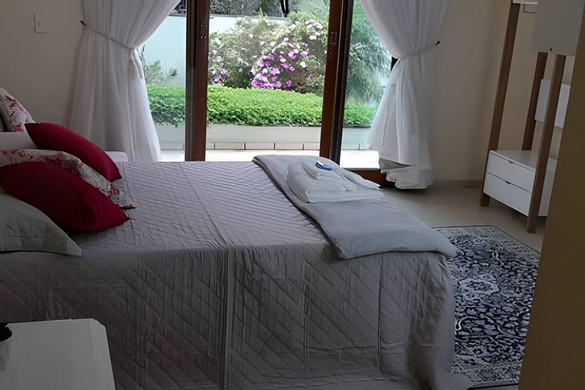 Quarto com cama, cortinas brancas, vista para um jardim e cômoda branca.