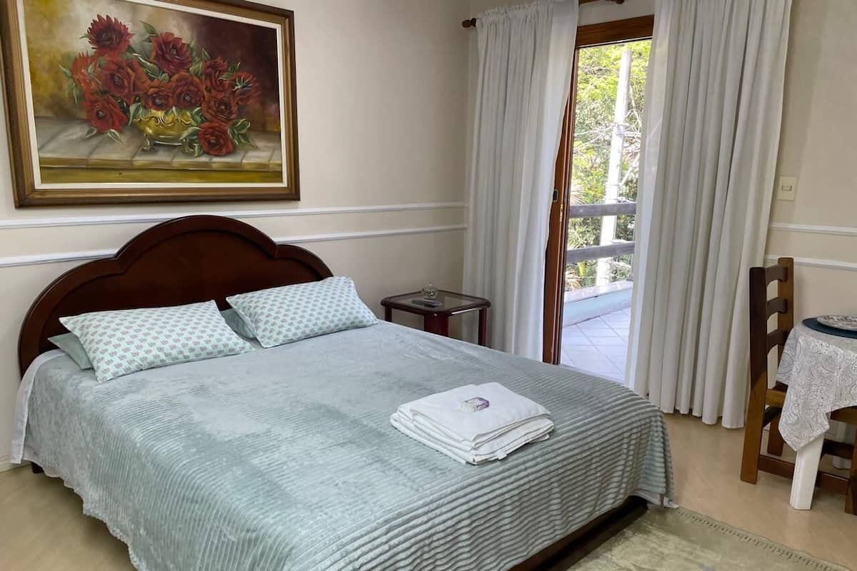 Uma cama bem arrumada com colcha estampada, uma mesinha de cabeceira com abajur e um quadro de flores na parede.