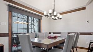 A dining area with a large rectangular table, six chairs, a chandelier, and a view through a large window. - Algonquin by RVMT (Mont-Tremblant)