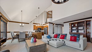 A spacious living area with a sofa, coffee table, and dining table. There is a kitchen in the background with wooden cabinets and a staircase. - Algonquin by RVMT (Mont-Tremblant)