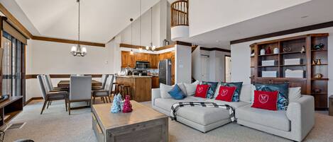 A spacious living area with a sofa, coffee table, and dining table. There is a kitchen in the background with wooden cabinets and a staircase.