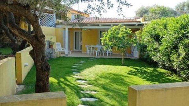 A yellow building with a tiled roof, a small patio with white chairs, and a well-maintained lawn with stepping stones. - Apartment in Sardinia near Surf Paradise (Porto Pollo)