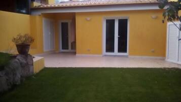 A yellow building with a tiled roof, a glass door, and a small garden area with a stone wall and a potted plant.