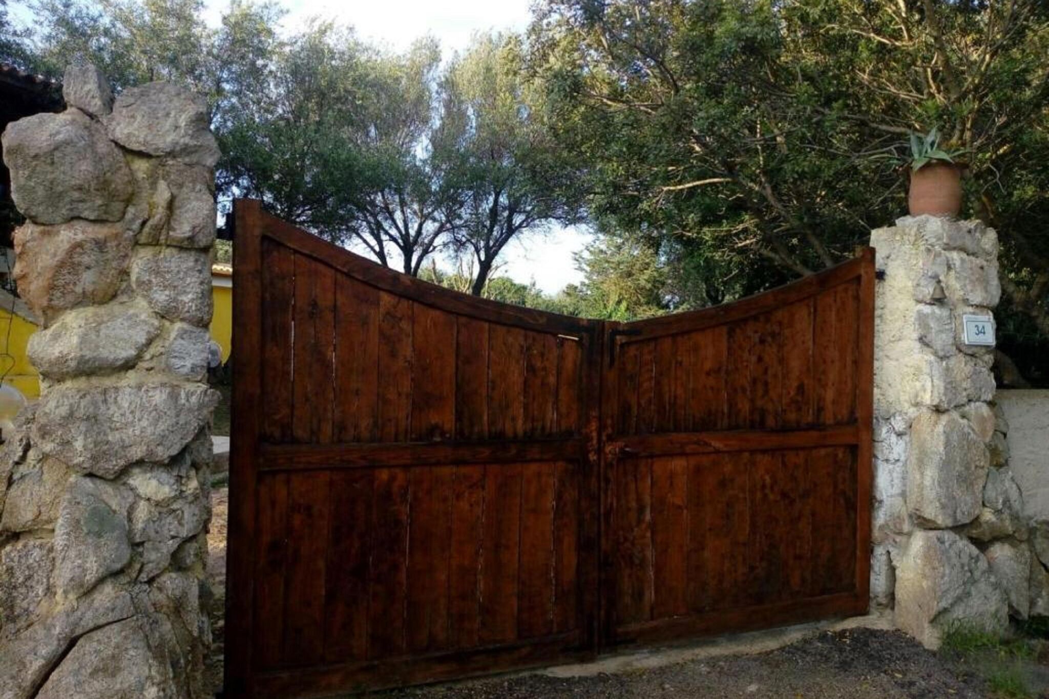 A rustic wooden gate with a stone wall on either side, leading to a property with a yellow building in the background.