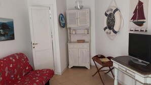 A room with a red patterned sofa, a white cabinet with shelves, a television on a wooden table, and a door. - Apartment in Sardinia near Surf Paradise (Porto Pollo)