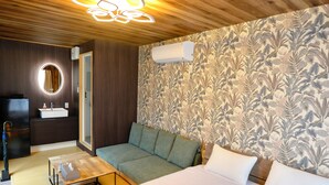 A modern hotel room with a wooden ceiling, a floral wallpaper, a sofa, a bed, a small table, and a bathroom with a mirror and sink. - Yonaguni-mumbu Airport Hotel (Yonaguni)