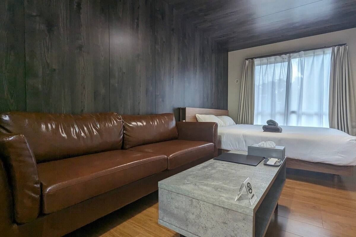 Ein modernes Hotelzimmer mit braunem Ledersofa, einem kleinen Couchtisch, einem Bett mit weißer Bettwäsche und einem großen Fenster mit Vorhängen.