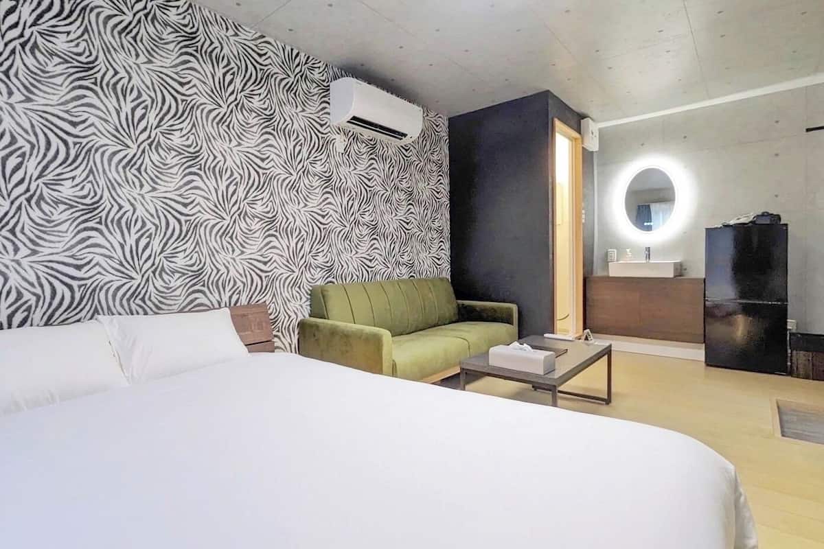 A hotel room with a bed, a green armchair, a small table, a mirror, and a black refrigerator.