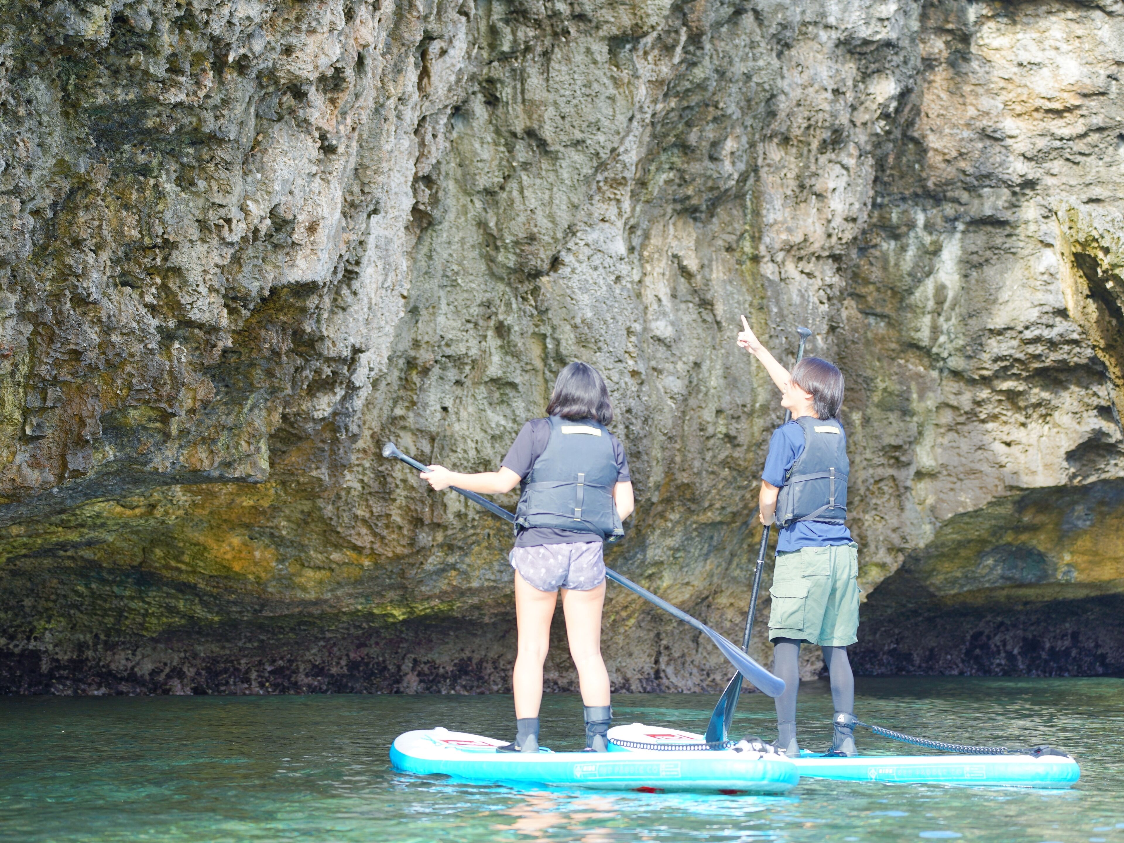 Deux personnes pratiquent le stand-up paddle sur une étendue d'eau claire, avec une falaise rocheuse en arrière-plan.