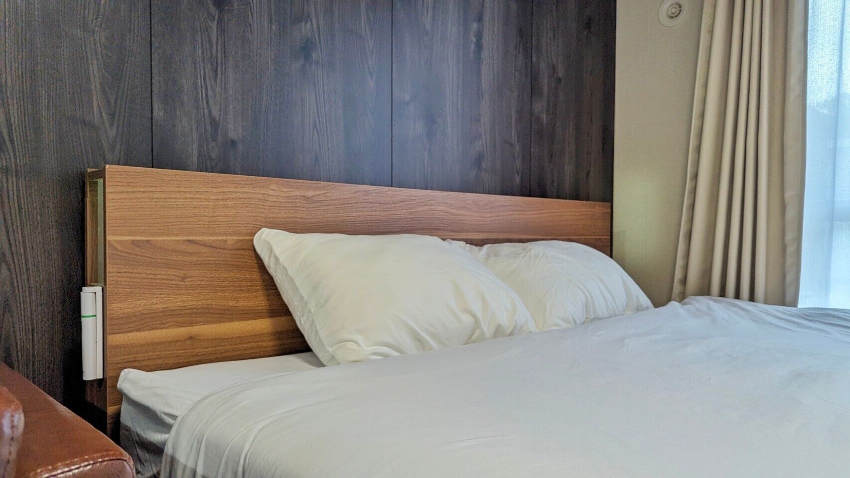 Una cama bien hecha con sábanas blancas y cabecera de madera en una habitación con paredes de madera oscura y una ventana con cortinas.
