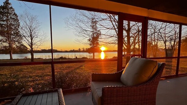 Une pièce avec vue sur un coucher de soleil se reflétant sur une étendue d’eau, une chaise en osier et une petite table.