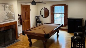 Une pièce spacieuse équipée d’une table de billard, d’un parquet, d’une cheminée et d’une grande fenêtre avec des volets.