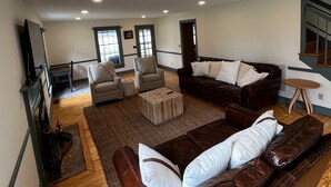 Un salon spacieux avec un canapé en cuir marron, un fauteuil beige et un canapé marron foncé. On y trouve une table basse en bois et une petite table ronde avec une chaise. Le sol est en bois et les murs sont blancs.