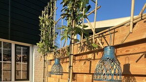 Una valla de madera con una planta de bambú en primer plano, un muro de ladrillo y una ventana con persianas.