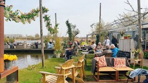 Un espacio al aire libre con sillas y mesas de madera, un cuerpo de agua al fondo y una estructura de invernadero.