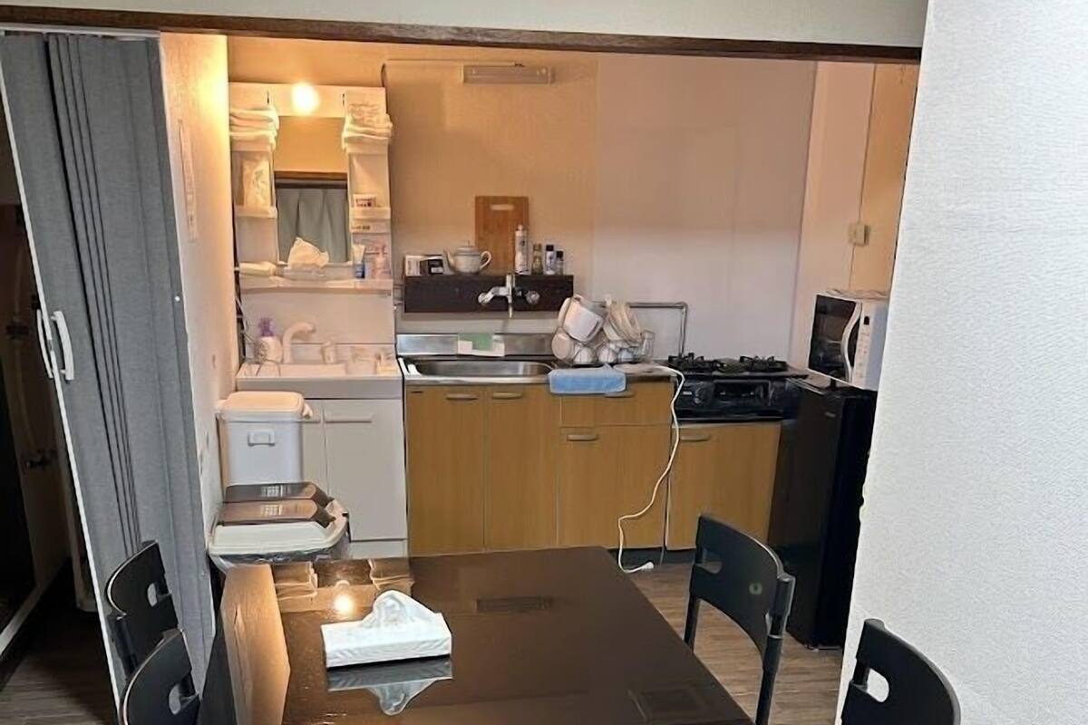 A compact dining area with a wooden table and four chairs, a kitchen in the background with a microwave and sink, and a small cabinet with toiletries.