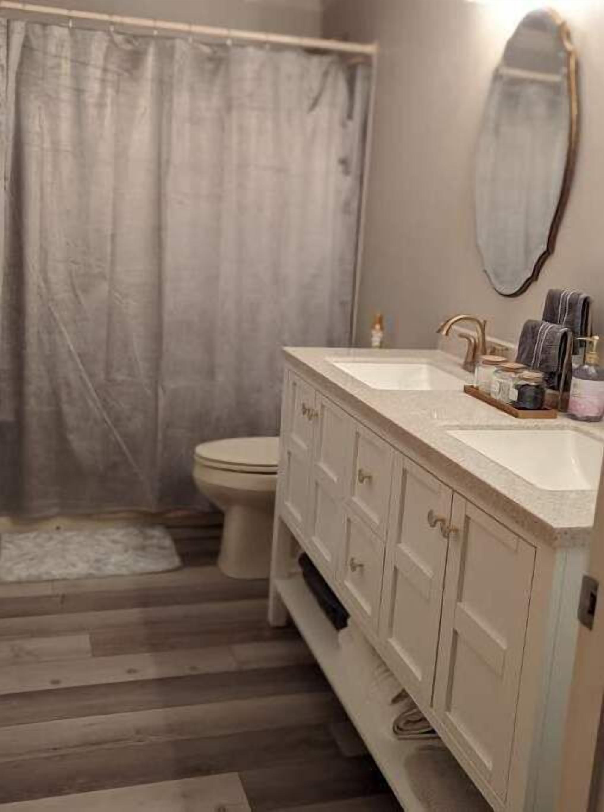 Un baño con piso de madera, lavamanos blanco, inodoro y cortina de ducha.