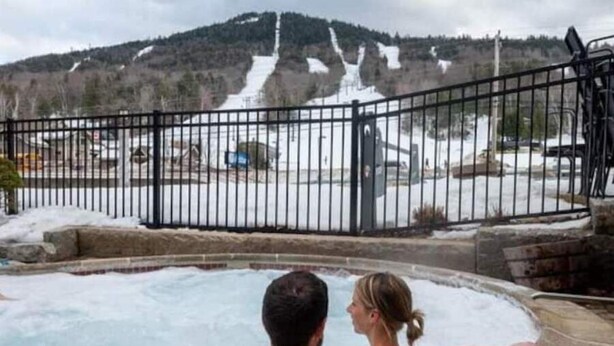 A couple relaxing in a hot tub with a view of a snowy mountain and a fenced area in the background.