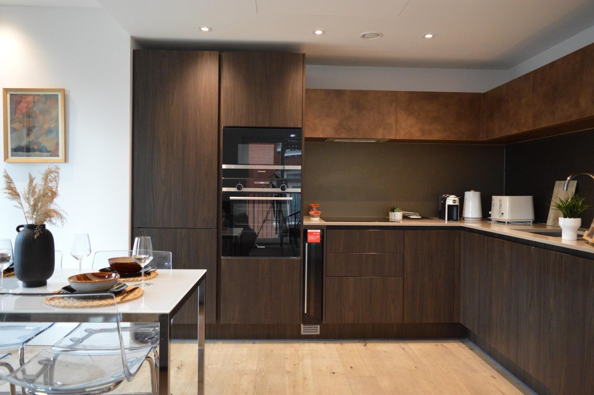 Een moderne keuken met donkerhouten kastjes, roestvrijstalen apparatuur en een glazen eettafel met een vaas gedroogde planten.
